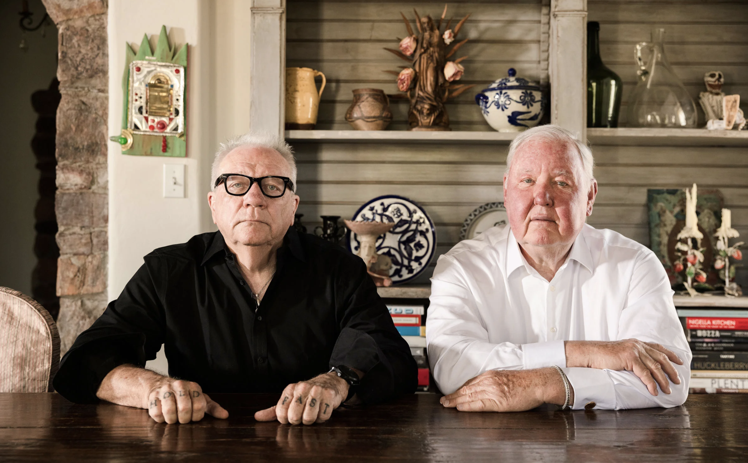 Two men sitting at a large wooden table with a shelf of books and knick knacks behind them.